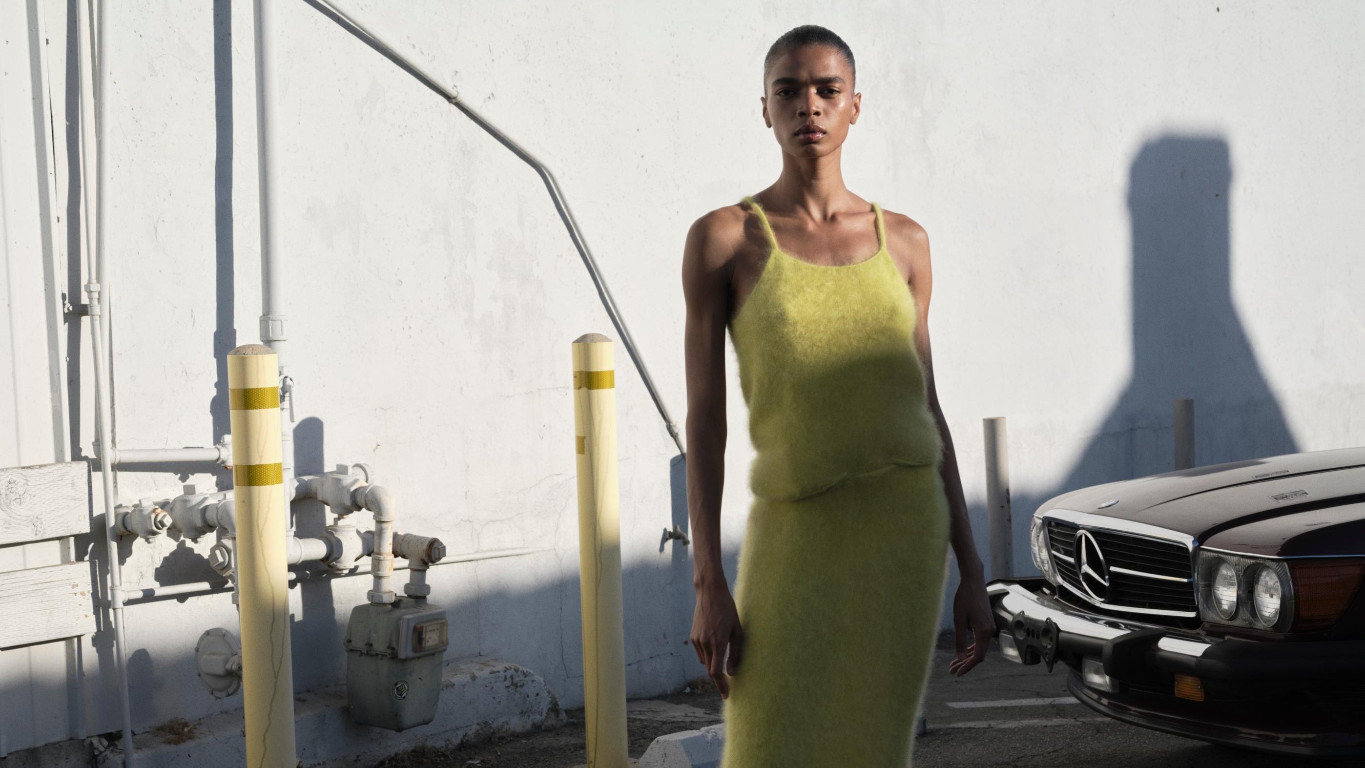 Model in lime brushed cashmere skirt and singlet top stands beside a black car in an outdoor fashion shoot.