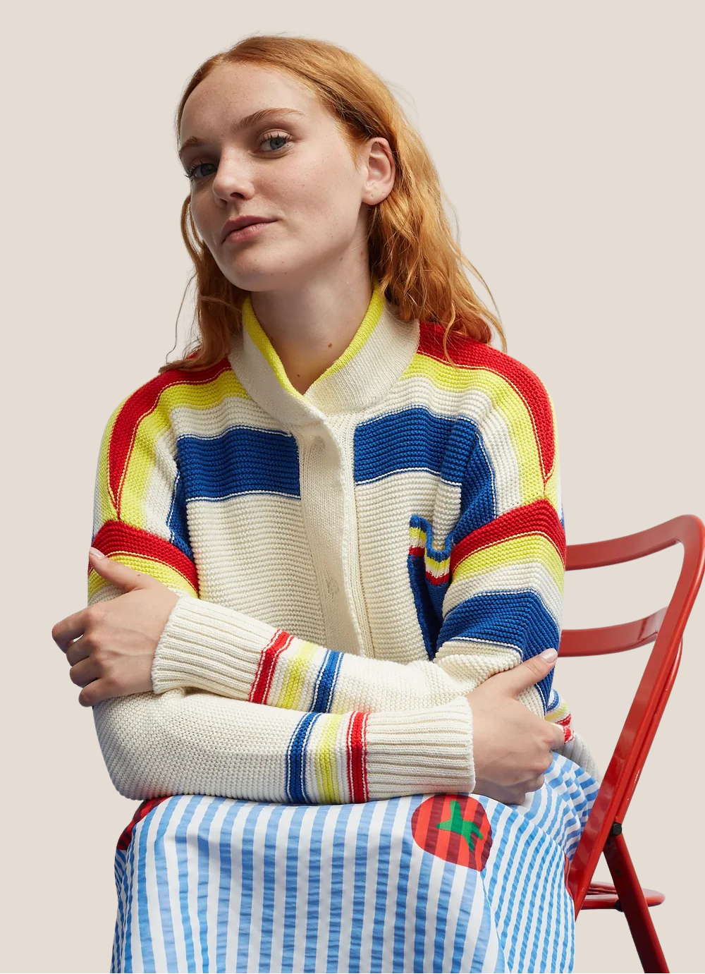 Model wearing the Dr Bloom Ecru Jersey Tennis Cardigan, seated on a red chair.